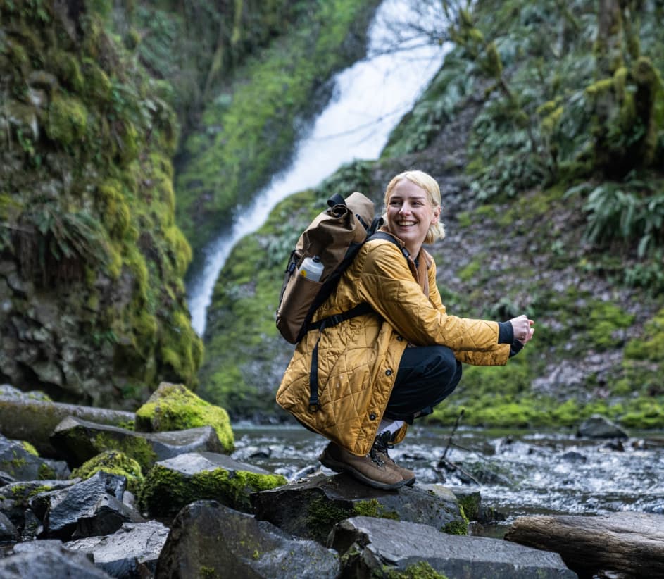 woman on a waterfall hike with a coyote daypack crouching by a river