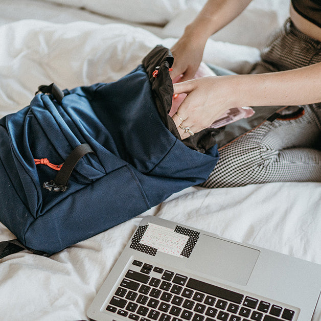 Woman packing her backpack on a bed with a laptop.