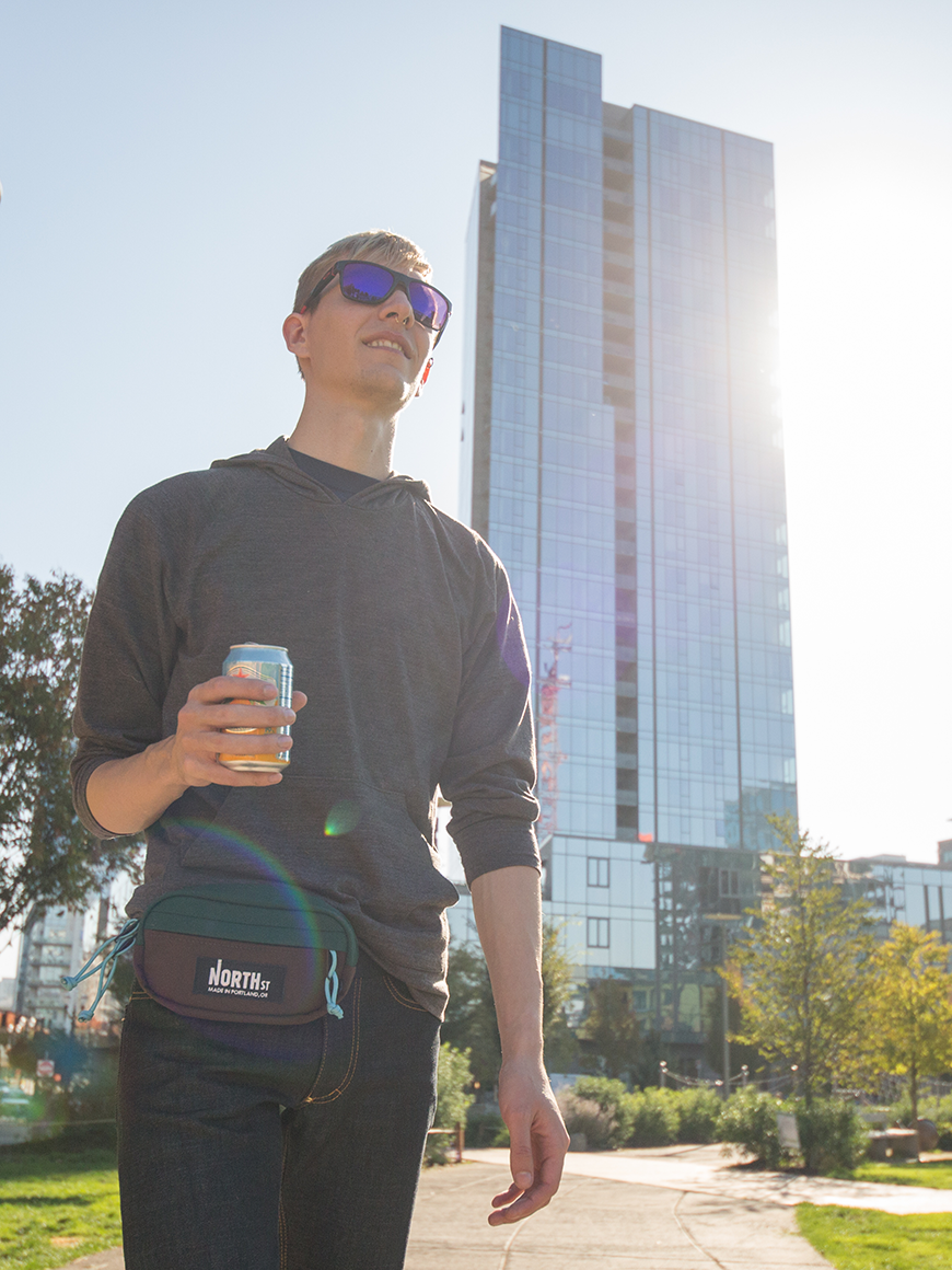 Young Man walking in city park with a cold drink wearing a hip pack - North St. Bags all-groups